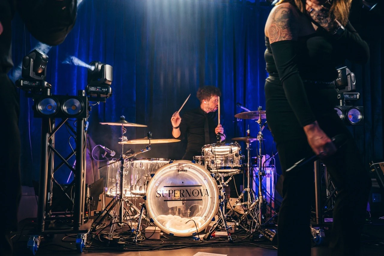 Callum drumming framed between vocalists with blue curtain backdrop